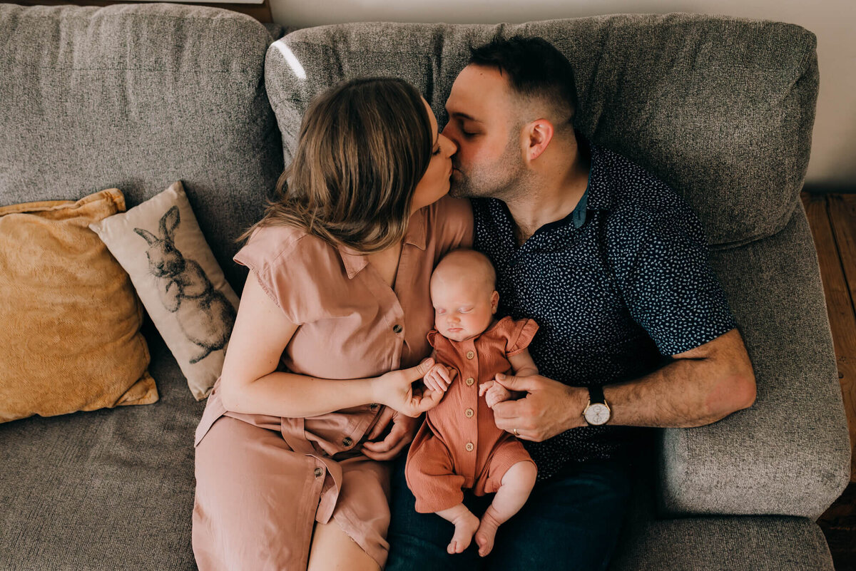 A young couple hold their newborn daughter on their couch while giving each other a kiss