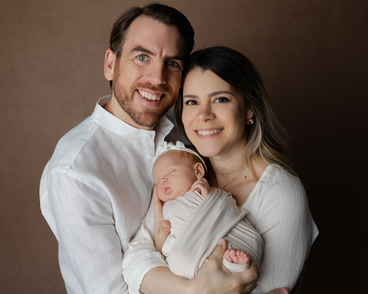 Heartwarming family portrait of proud new parents holding their newborn baby girl, wrapped in soft cream tones. Photographed in-studio by Inland Empire newborn photographer Kaitlyn Dawn Photography.
