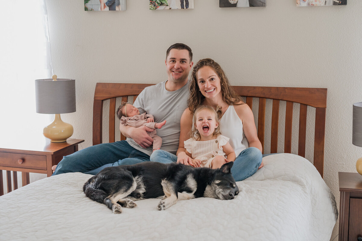 Happy picture of a family of four and their dog sitting on their bed and smiling  at the camera.