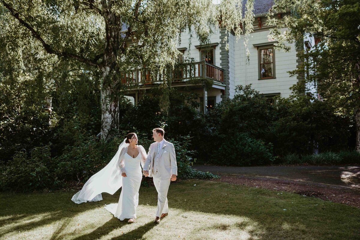 A Bride and Groom hold hands and walk through a patch of sun in front of an old white manor in Oregon