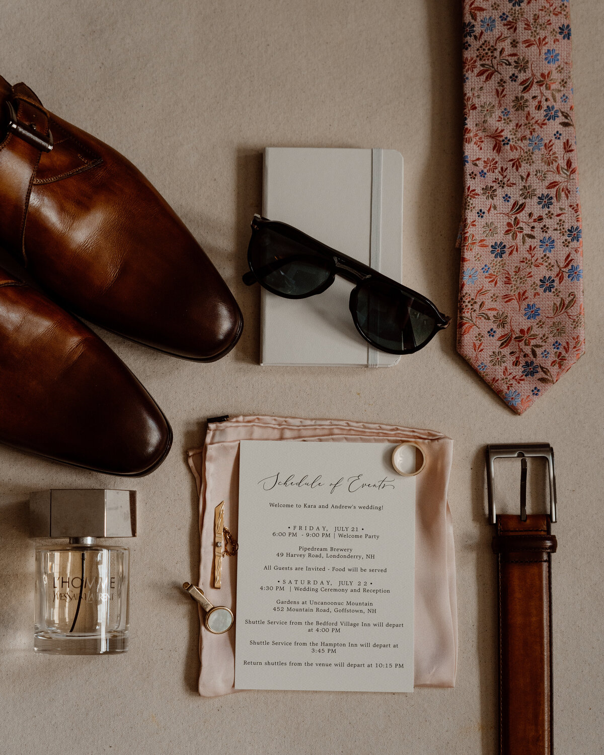 Flat lay of groom's wedding day essentials including brown leather shoes, floral tie, sunglasses, cologne, belt, schedule of events, and accessories