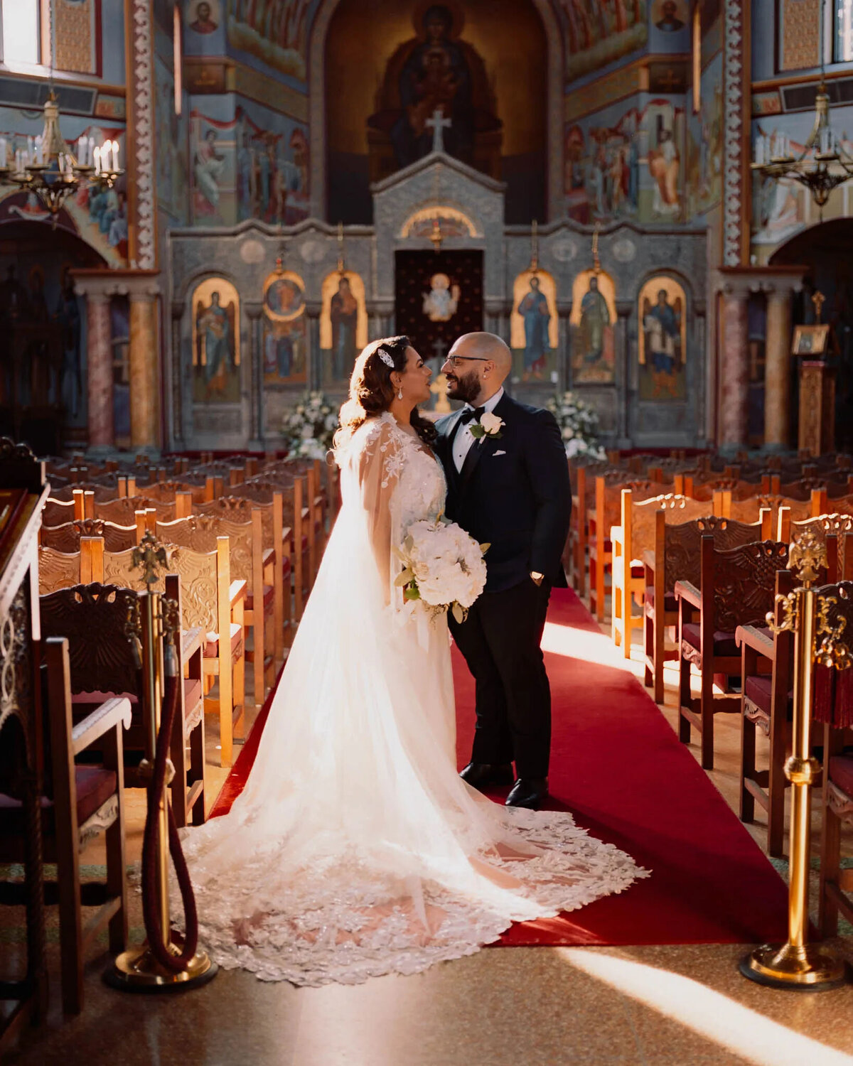 A bride and groom stand together on a red carpet in a beautifully decorated church, gazing at each other and smiling. Captured by an NJ wedding photographer, the moment glows as sunlight streams in and the bride holds her bouquet.