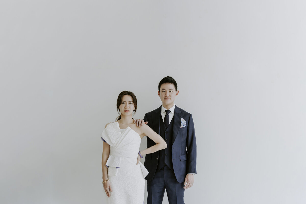 the groom putting her hand at the bride's shoulder while posing for the camera with white wall as their backdrop