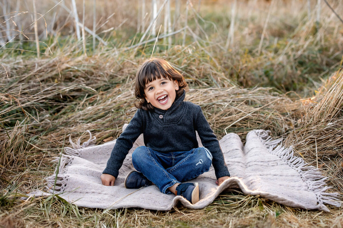 A joyful child with shoulder-length hair sits on a fringed blanket in a grassy field, wearing a dark sweater and jeans. The child is smiling with excitement, surrounded by dry grass and plants—an ideal moment for outdoor portraits captured expertly by a Bloomington family photographer.