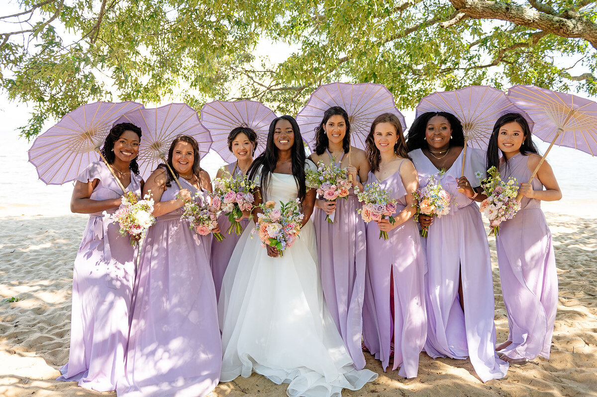 Bride and bridesmaids dressed in lilac gowns holding pastel floral bouquets, posing under matching lilac umbrellas. at the Chesapeake bay  foundation.