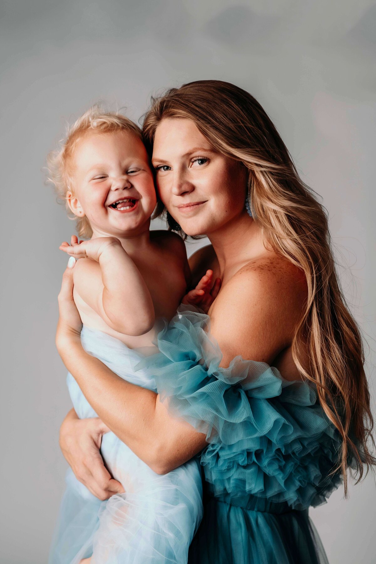 A close-up photo shows a young mom and her toddler daughter, both in matching blue tulle. The child looks very happy and playful as she grins and waves at the camera, while her mom holds her against her body and snuzzles in close for an intimate, cuddly, motherhood portrait by Mobile, AL Motherhood Photographer