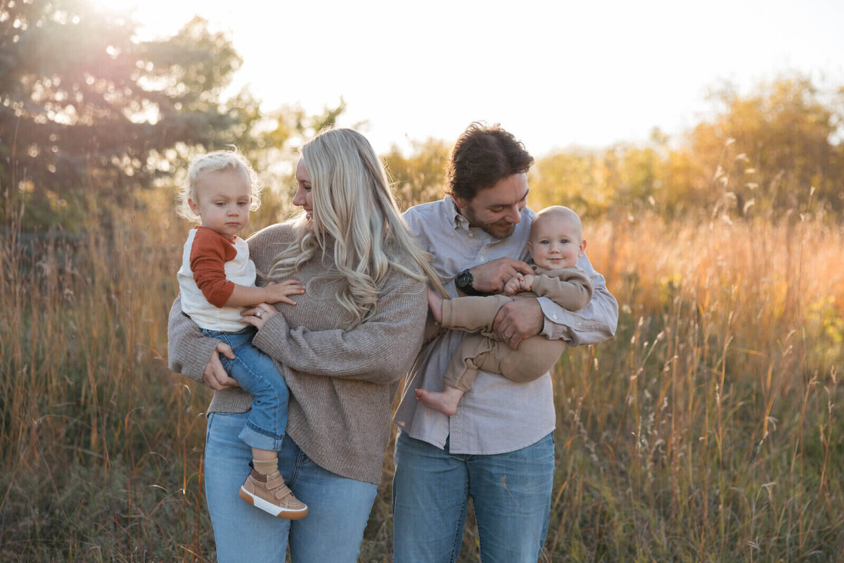 A young family of four enjoying a golden hour photoshoot in a grassy field, with the mother holding a toddler and the father cradling a smiling baby.