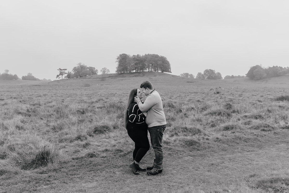 Couple standing closely forehead to forehead in the countryside with deer behind them