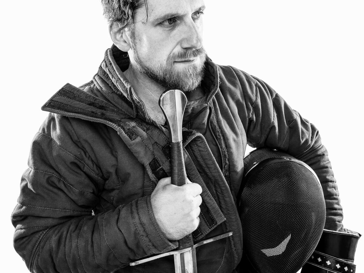 Studio editorial portrait of a male fencing athlete in full gear, holding his helmet and sword, captured in Austin, TX.
