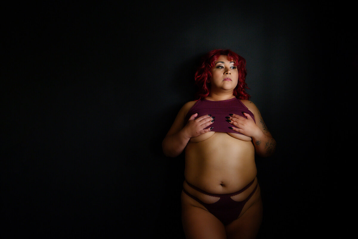 red-haired woman leaning against a lack wall wearing burgundy crop top and underwear.