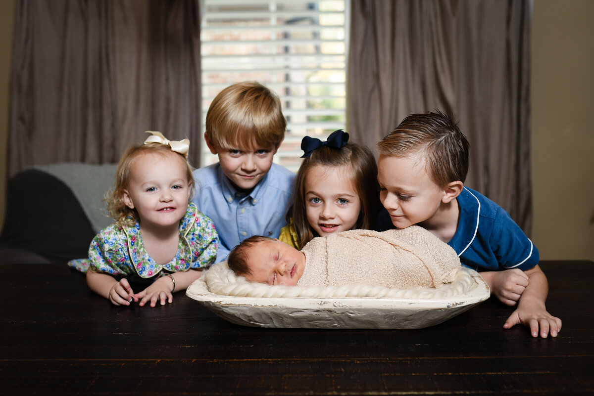 Beautiful lifestyle newborn photography: Siblings look over newborn baby in dough bowl on family dining table in Madison, MS
