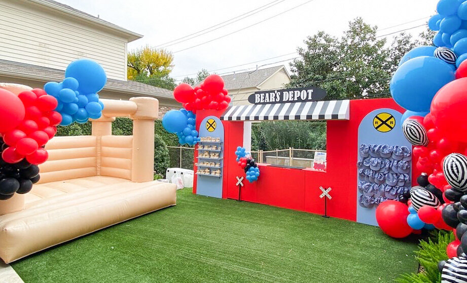 Kids train depot themed birthday backdrop display with blue and red balloons and a tan inflatable bouncy house