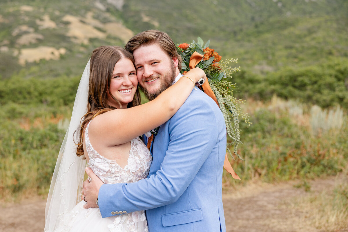 The bride and groom share a joyful embrace, smiling at the camera. The bride’s veil cascades down her back as she rests her arms around her groom’s shoulders, holding her bouquet behind him.
