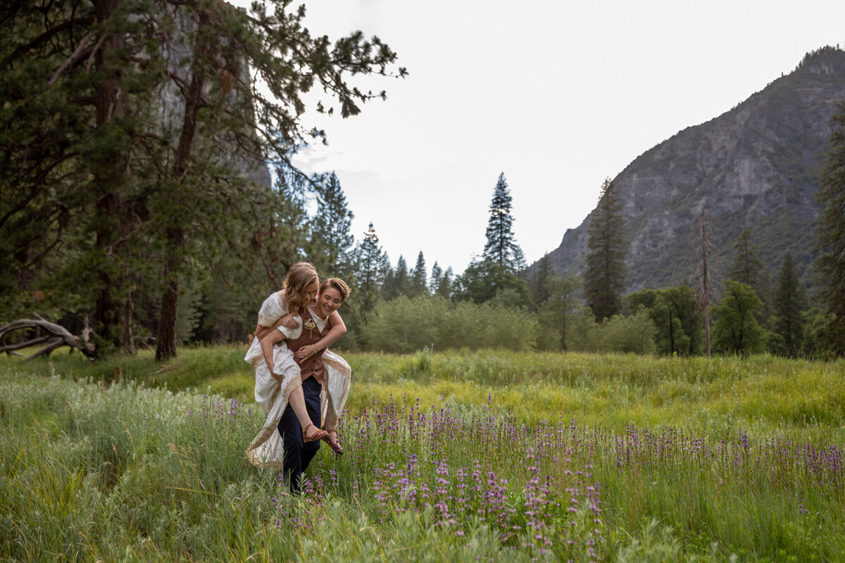 A bride gives her new wife a piggy back ride as they walk through a field of lupin flowers in Yosemite.