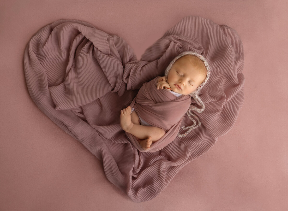 Brooklyn newborn photoshoot. Newborn baby girl is swaddled in mauve with her legs and fingers peeking out of the wrap. Baby is placed on ruched fabric in the shape of a heart. Captured by top Brooklyn newborn photographer Chaya Bornstein