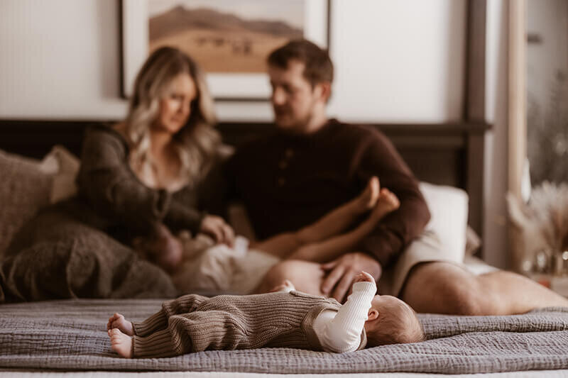 Mom and dad tickle toddler daughter in the background while newborn baby son sleeps at the foot of the bed in focus
