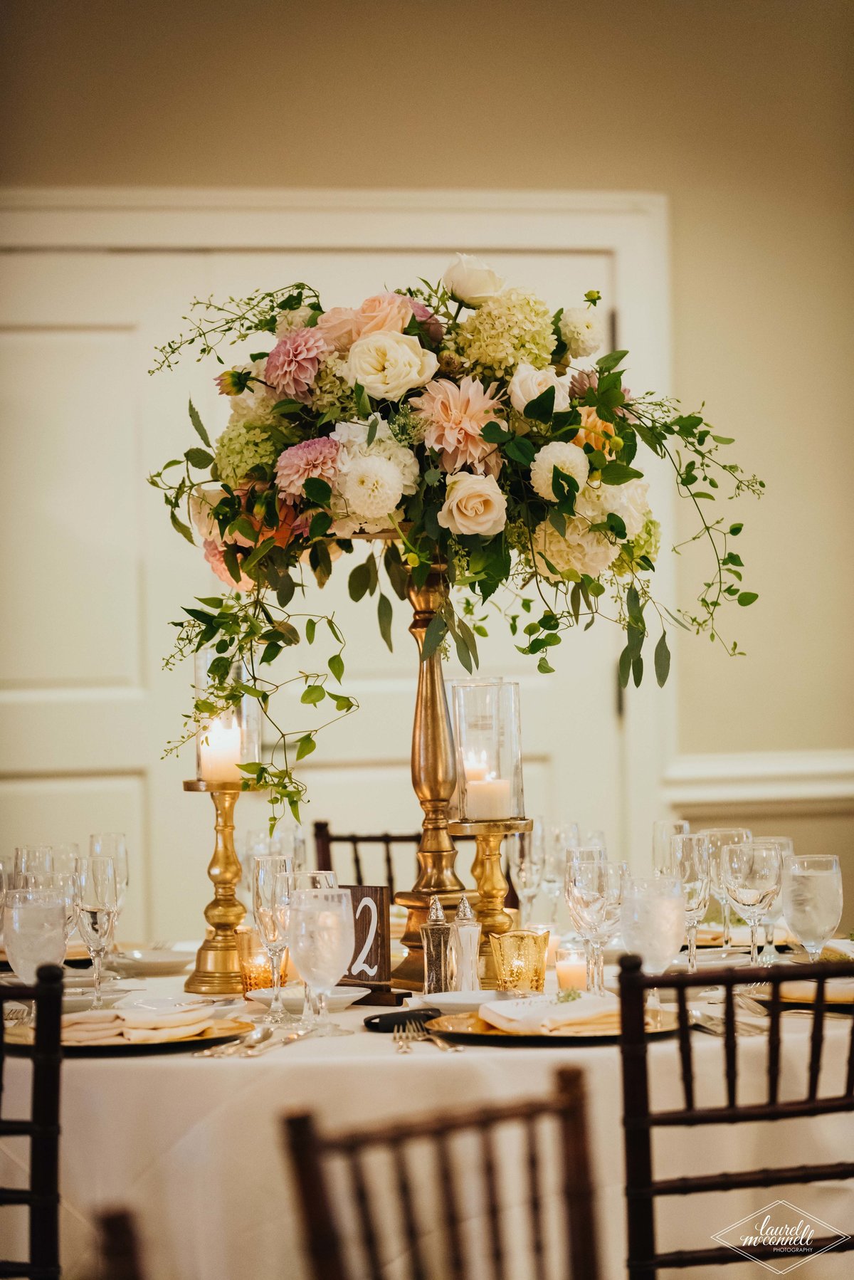 elevated centerpiece of cafe au lait dahlias, peach and ivory roses, smilax greenery, and raspberry foliage on table with ivory linens, gold candle holders, and brown Chivari chairs
