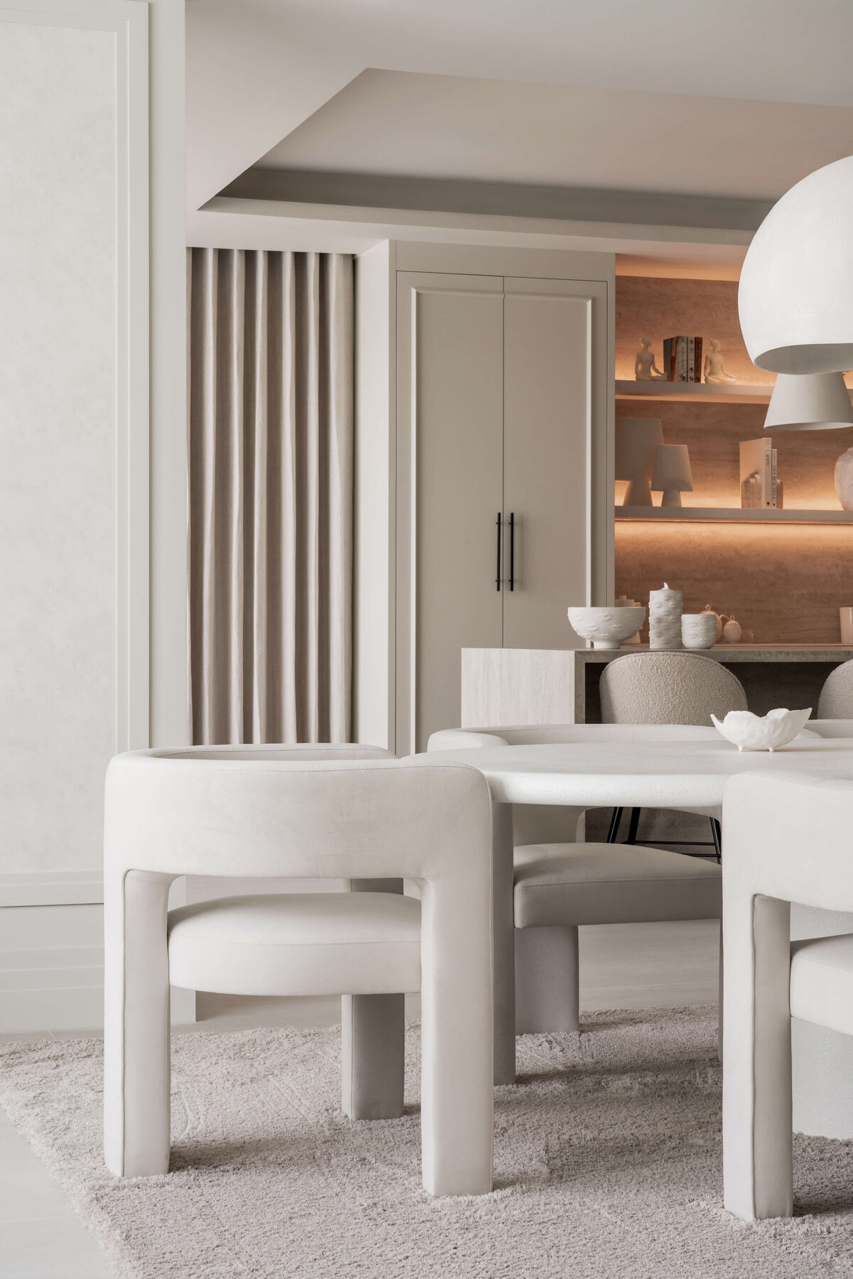 A modern dining room with a minimalist aesthetic, featuring a large oval table surrounded by curved white chairs. Close up on one chair at the head of the table. Behind, part of the kitchen can be seen with built-in shelving that is illuminated by soft, integrated lighting.