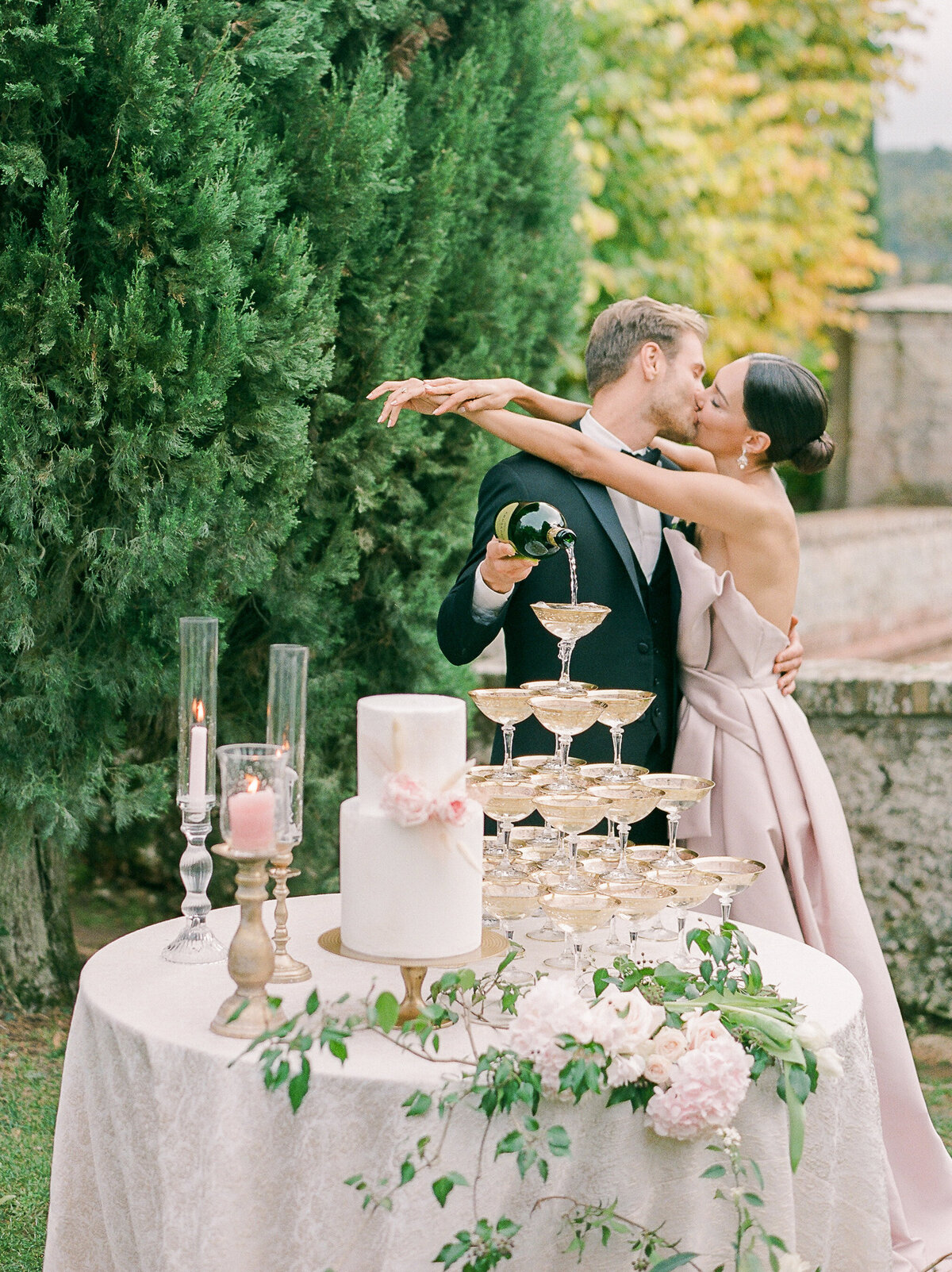 bride in a pink wedding dress with her arms around her groom while he pours champagne into the chamoagne tower that is on their wedding cake table in cetinal in tuscany italy