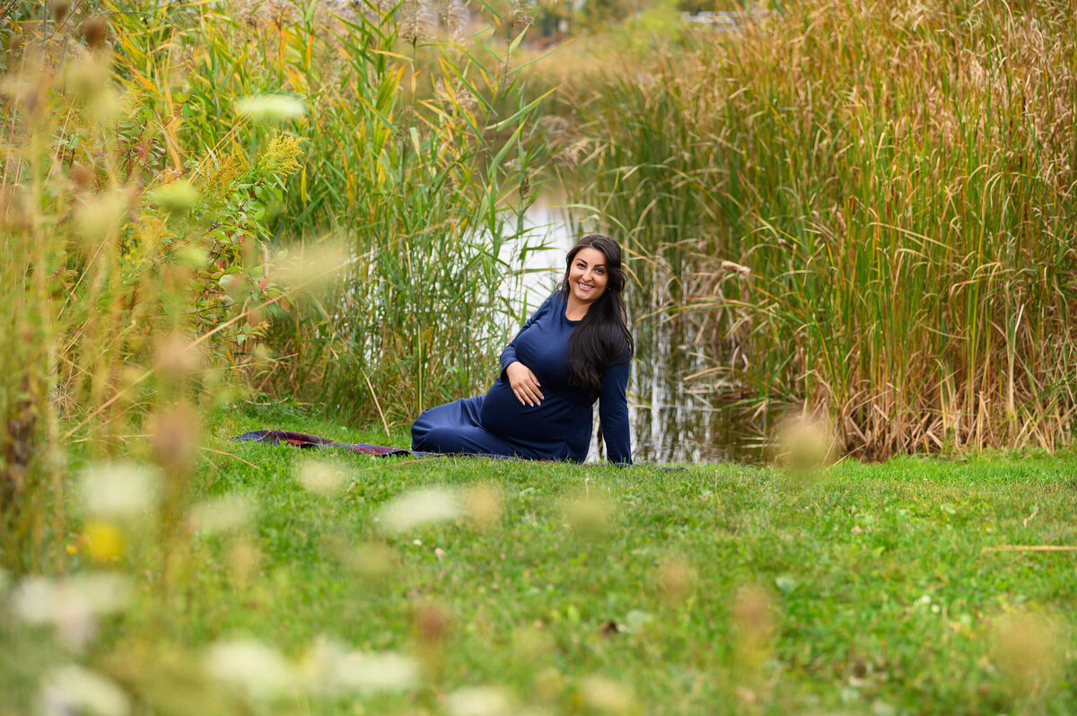 brown-haired pregnant woman sitting on the ground in front of a pond wearing a blue maternity dress for her outdoor fall Toronto Maternity photos.