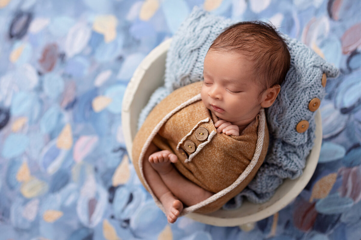 Newborn baby boy wrapped in a warm yellow swaddle, sleeping in a round bowl with blue knits on a soft blue painted backdrop.