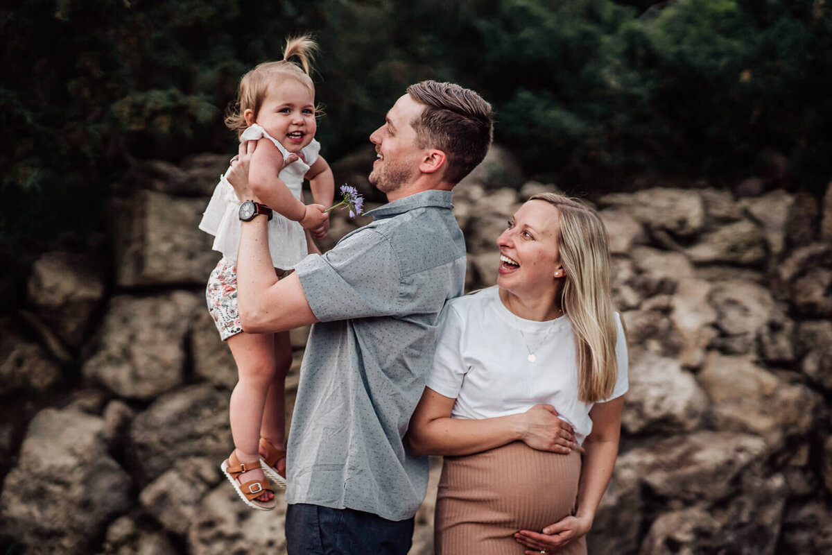 A father holds up his smiling toddler, while his pregnant wife stands back to back with him, smiling at her daughter.