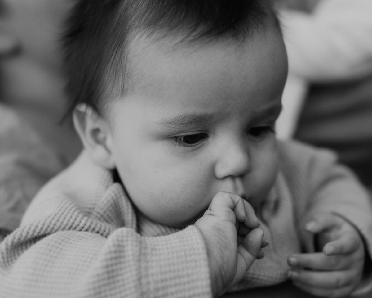 A black and white close-up of an adorable baby boy with a full head of hair, chubby cheeks and fingers in his mouth.