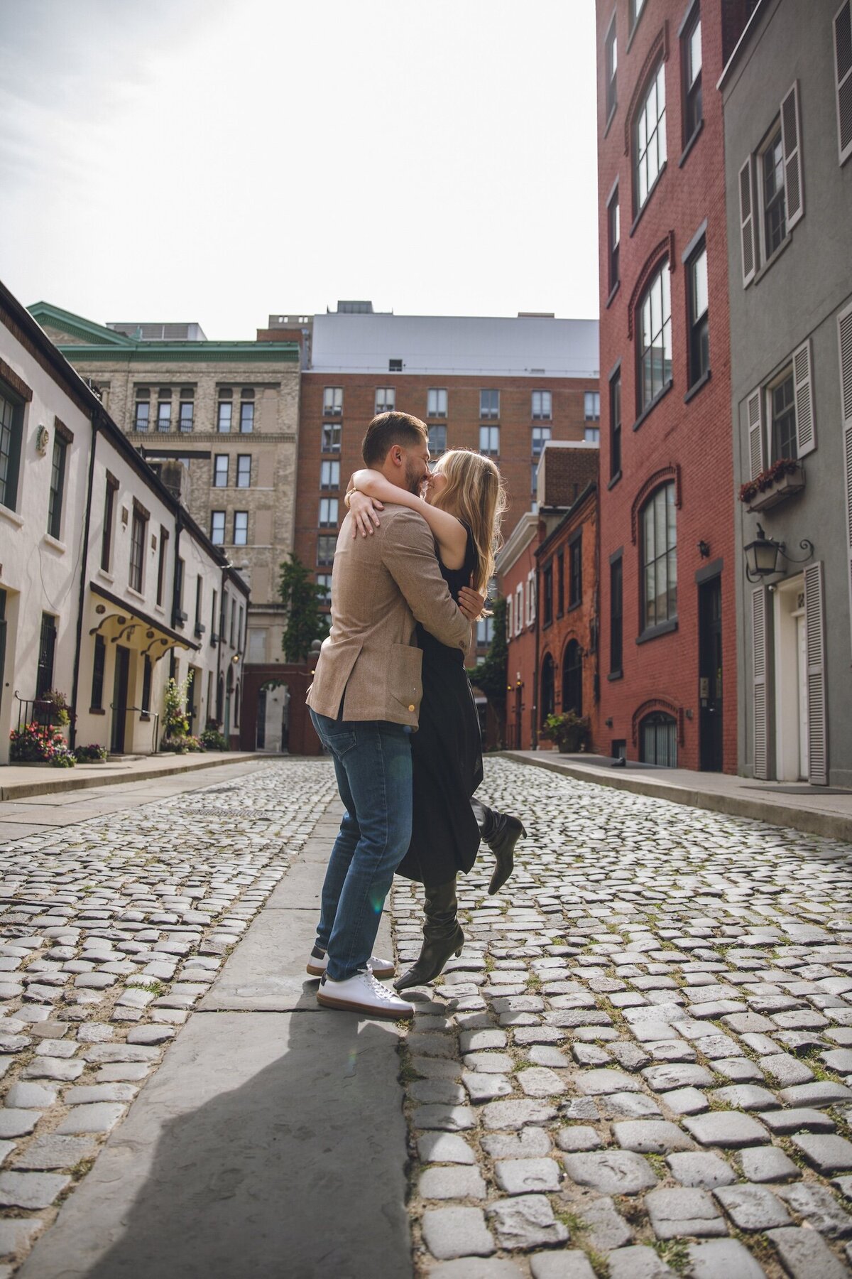 Groom lifting fiancée for a joyful kiss on the cobblestone street.