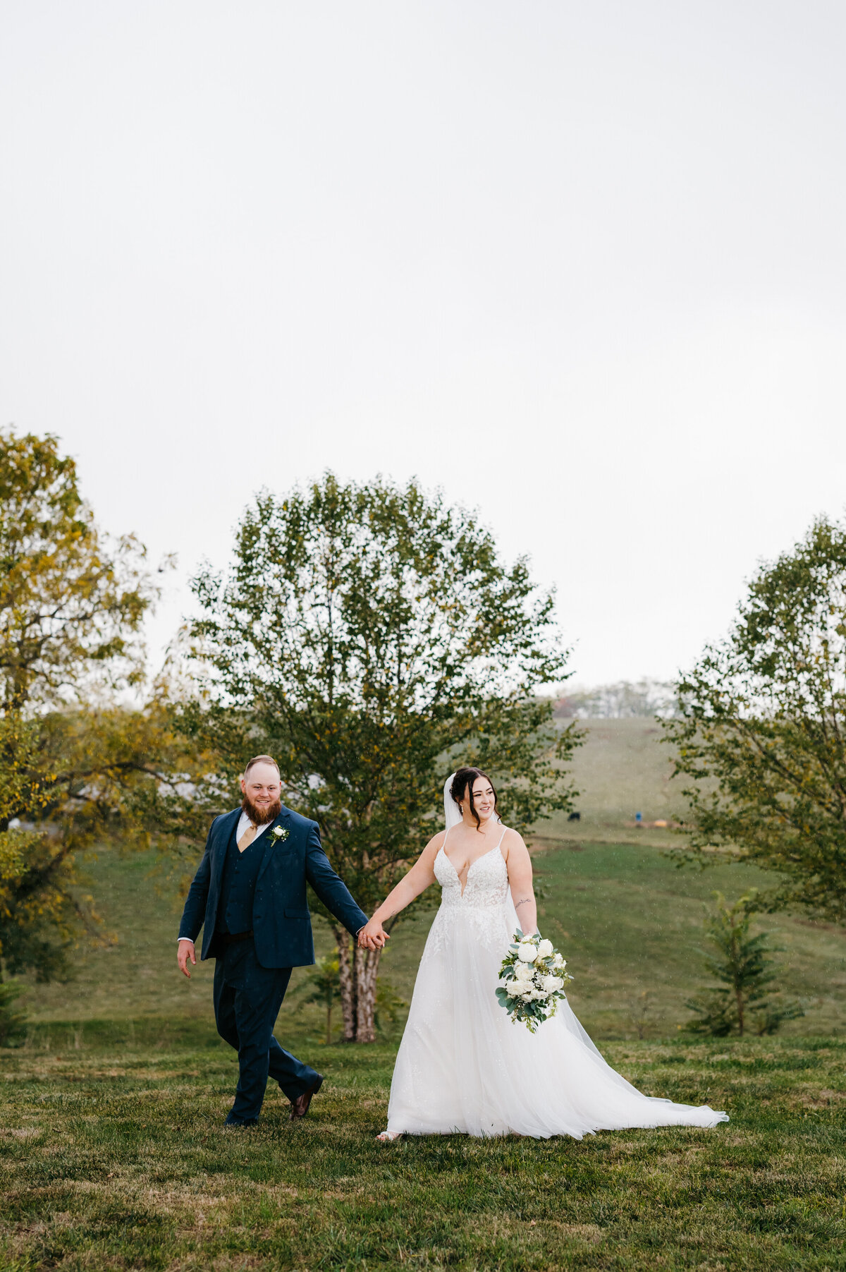 Sunny Slope Farm wedding with bride and groom holding hands and walking through a pasture together with trees in the distance and the sun shining over the mountain and onto the tops of those trees