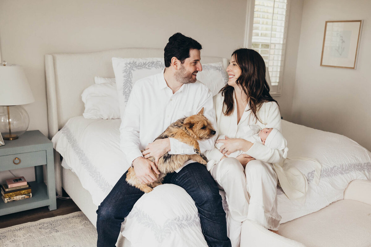 a father and mother sit on their bed while holding their dog  and newborn baby