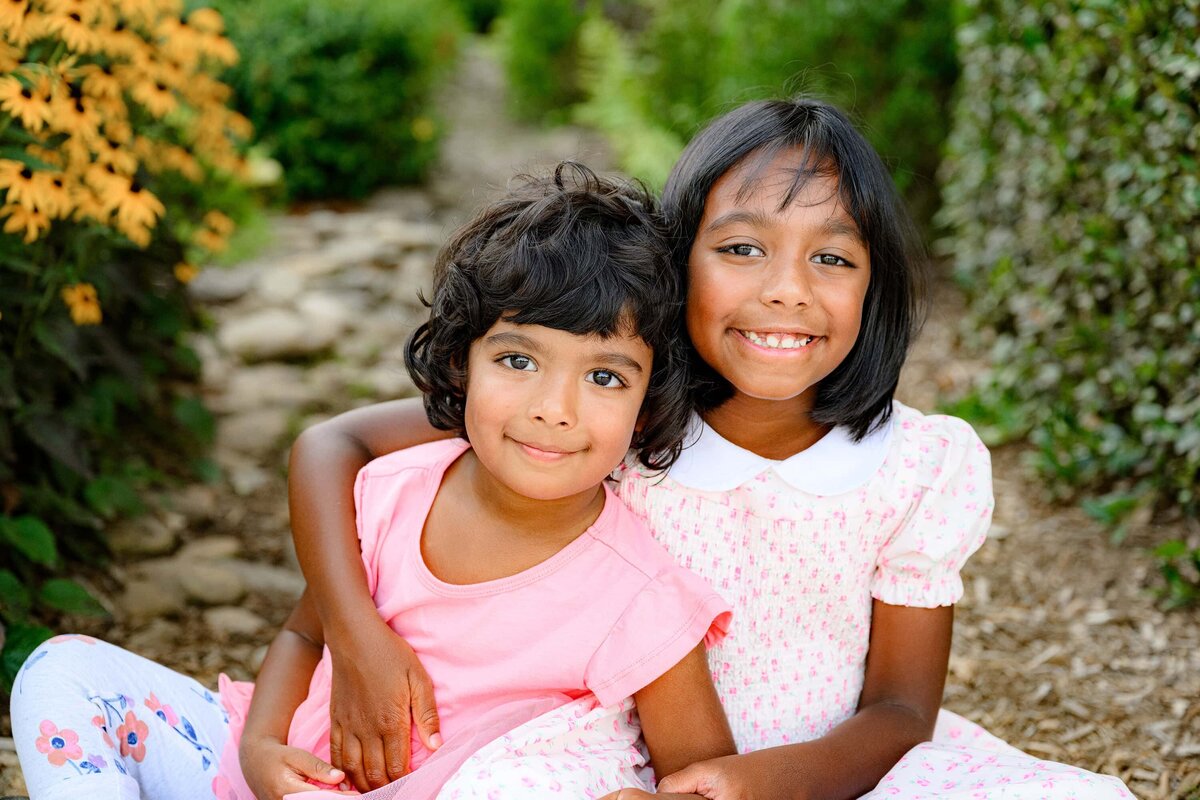sisters sit in a flower bed dressed in pinks and peaches with flowers behind them