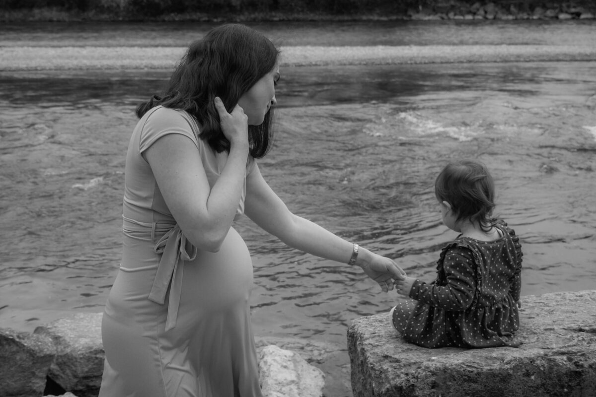 Pregnant woman holds the hand of her little daughter who watches the lake behind.