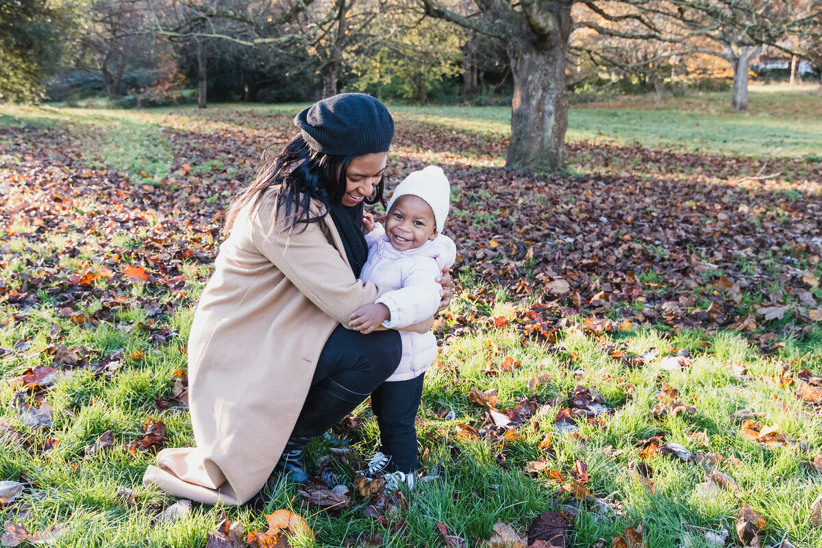 A smiling woman in a coat hugs a toddler in a park filled with fallen leaves.