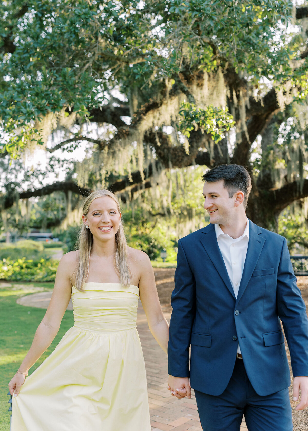 an engaged couple holding hands while walking among hanging moss trees
