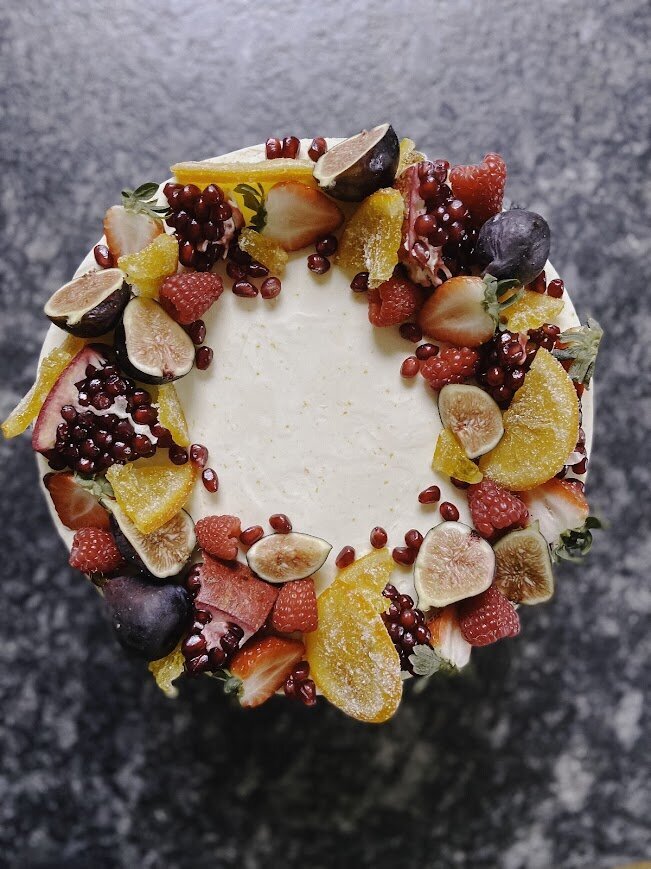 A top shot of a round cake with a wreath of fruits in warm tones - candied oranges, fresh figs, and pomegranate. 