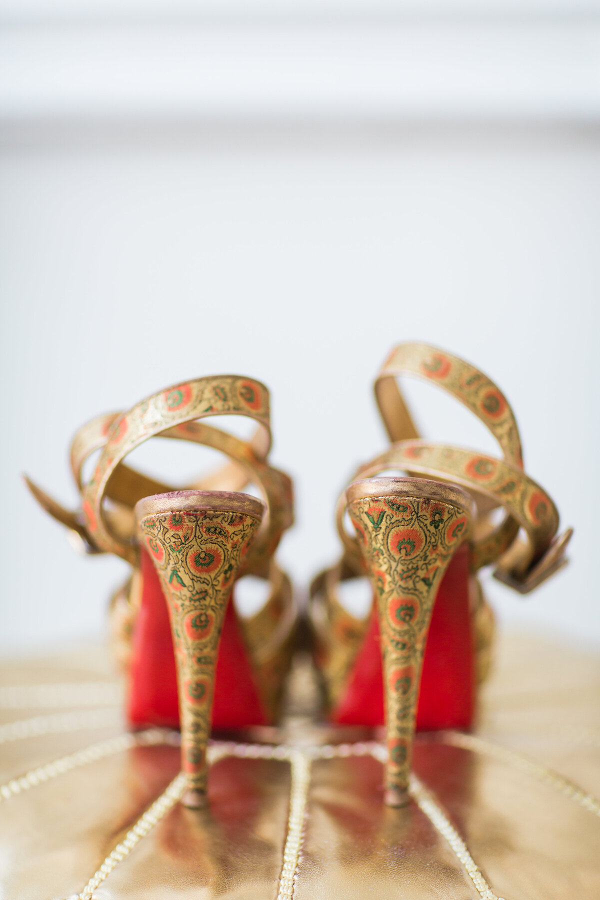 The photo was taken at the Mellon Auditorium in Washington, D.C., one of the best luxury venues in the city with European flair, captured by the talented photographer Sarah Bradshaw.   In the image, we see a close-up of a pair of ornate high-heeled shoes with intricate golden patterns and striking red soles. These exquisite shoes feature buckled straps and are elegantly positioned with the heels facing the camera, highlighting their detailed design and impeccable craftsmanship.
