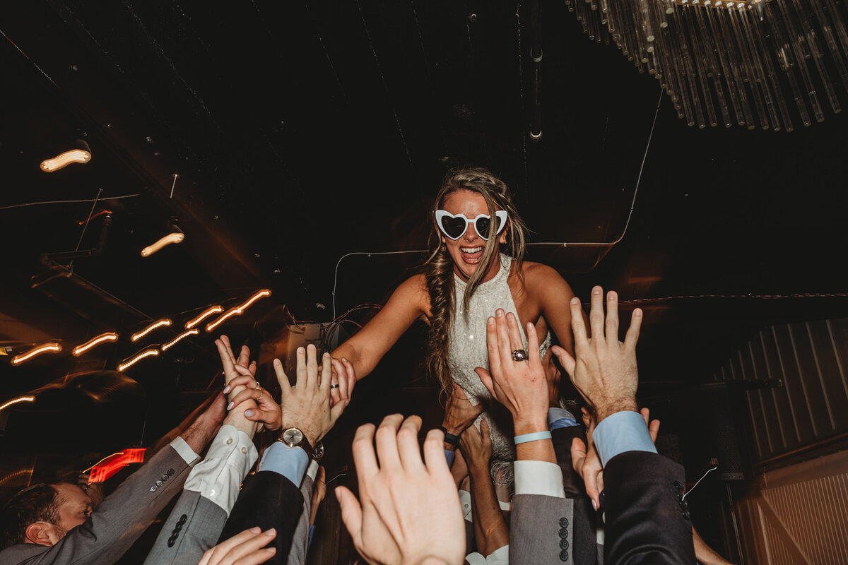 Maryland wedding photographer captures reception fun with bride being carried in a crowd surf while wearing heart glasses