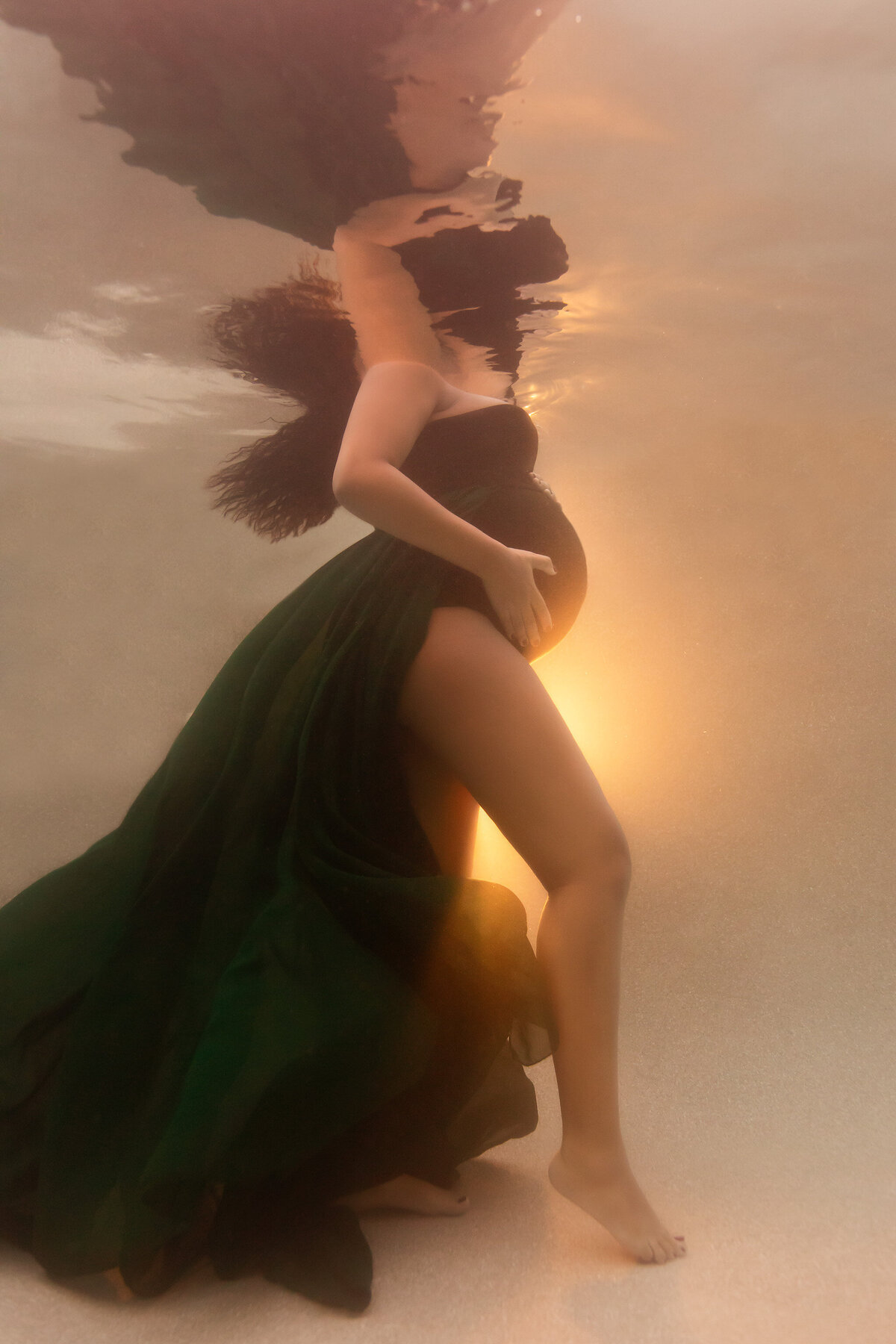 A captivating underwater maternity portrait featuring a woman in a flowing dark green gown, softly illuminated by a warm, golden glow. The tranquil setting highlights the ethereal beauty of the moment.