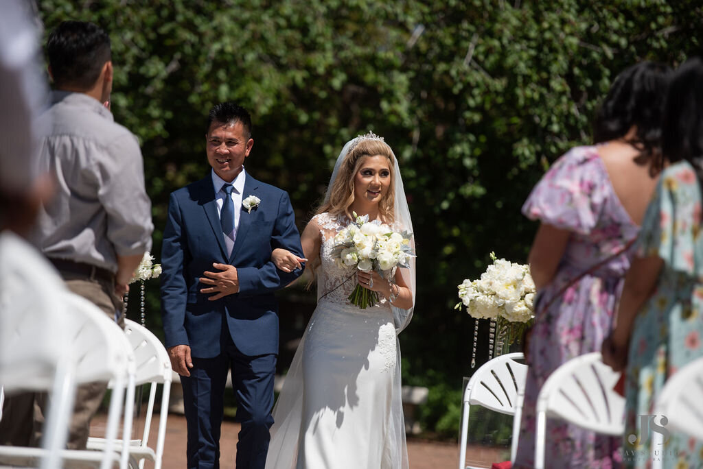 Bride walks down the aisle  with her father with a white bouquet.