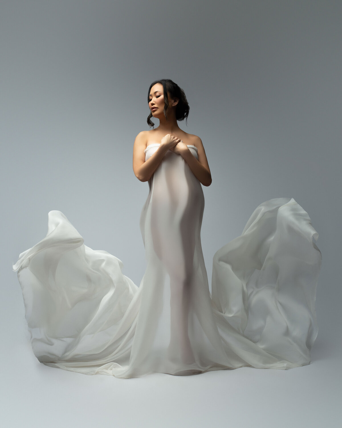 30 weeks pregnant stunning asian woman wrapped in flying white fabrics, closing her eyes with an all white backdrop in studio