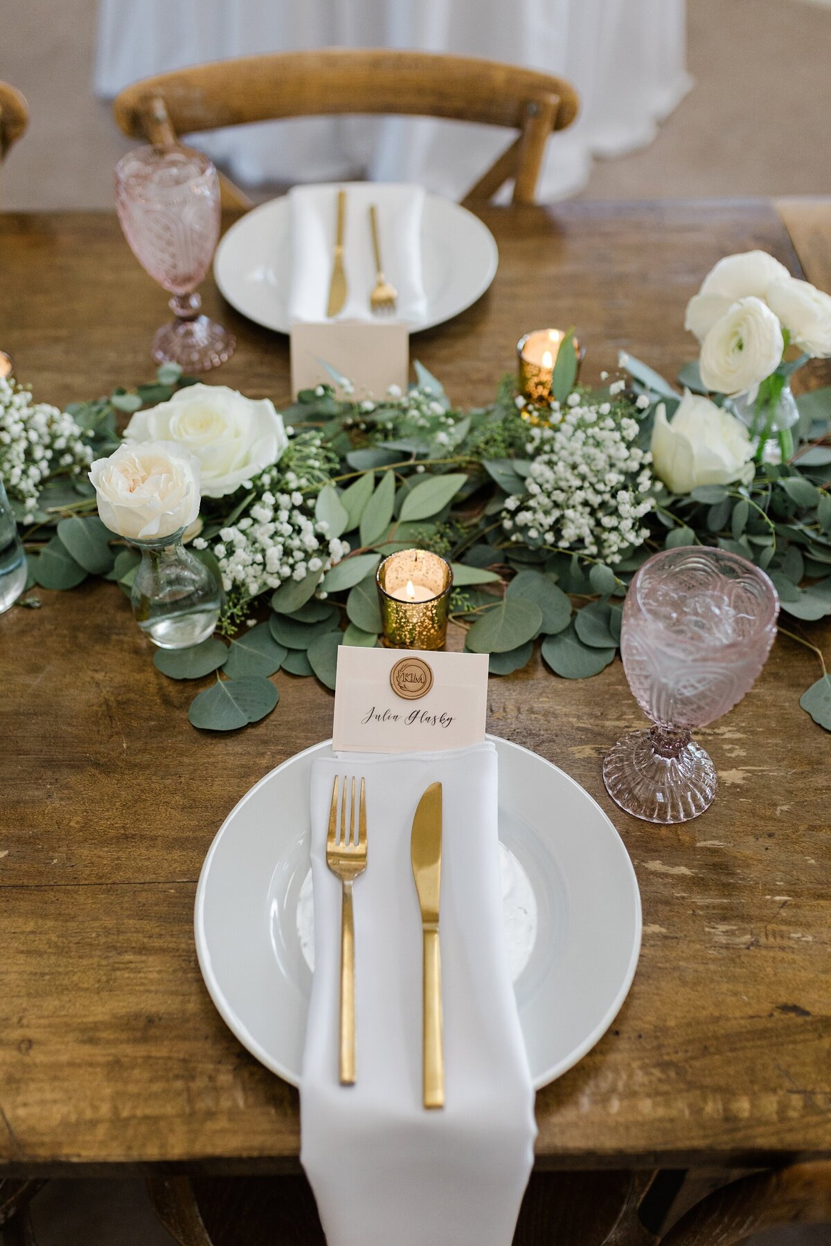A detail shot of a place setting for a wedding reception at Bella Cavalli Events in Aubrey, Texas. The place setting is made up of a white plate, gold knife and fork, napkin, and a personalized name tag embossed with a wax seal. Behind it is a candle and a long floral arrangement extending to either side. An identical place setting can be seen on the opposite side of the table.