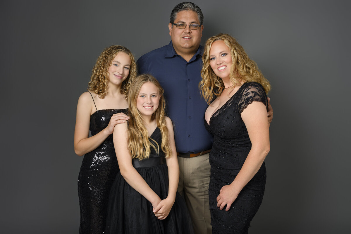 A family of four, with three females dressed in black, posing together against a gray backdrop