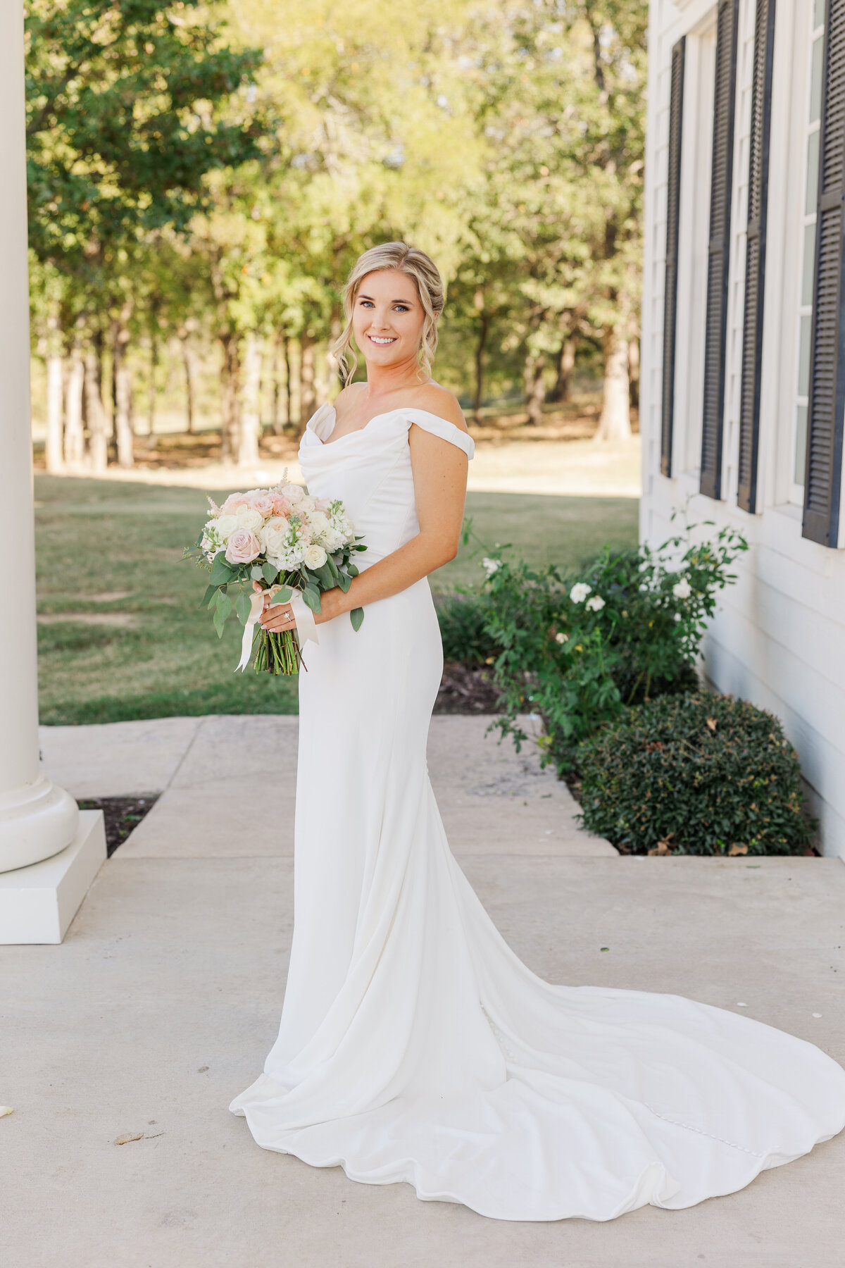 Radiant bride in a classic off-the-shoulder gown poses with blush and ivory bouquet outside a historic wedding venue near Flower Mound, TX. A timeless bridal moment full of grace and charm.