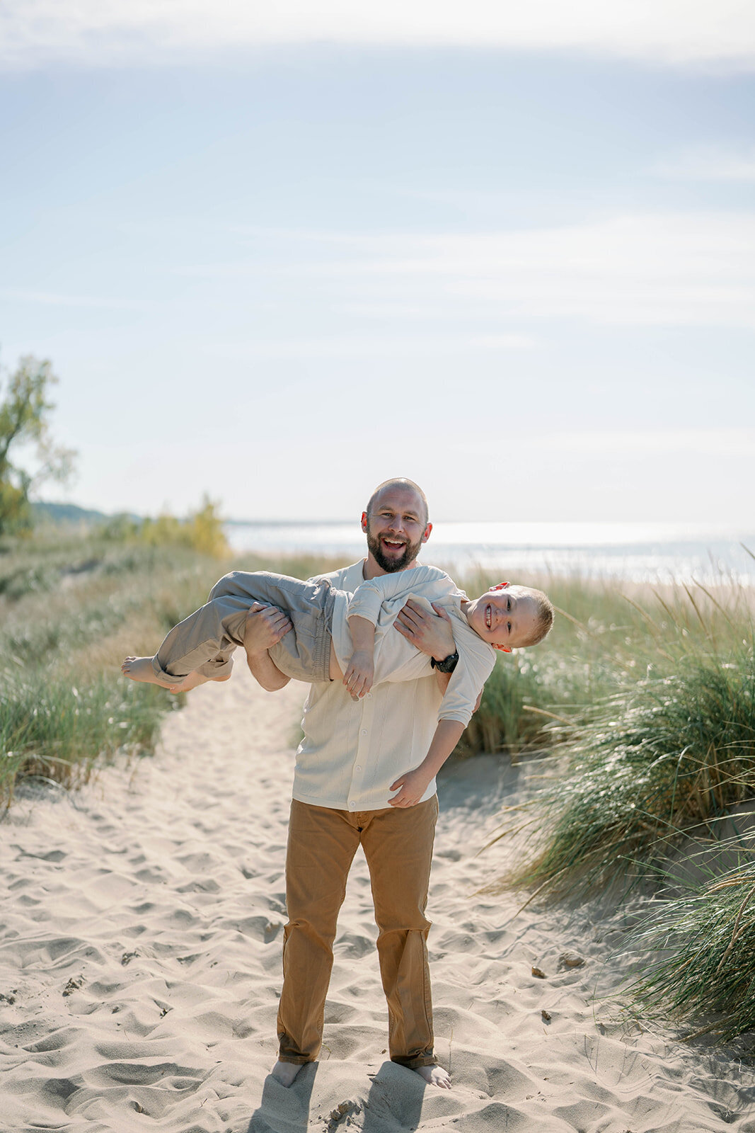 A father lifting his young son and laughing together on the sandy path at Weko Beach in Bridgman Michigan.