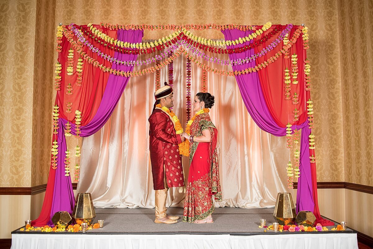 Hindu groom and bride wearing red and gold holding hands under a red and magenta mandap draped with strands of hanging carnations