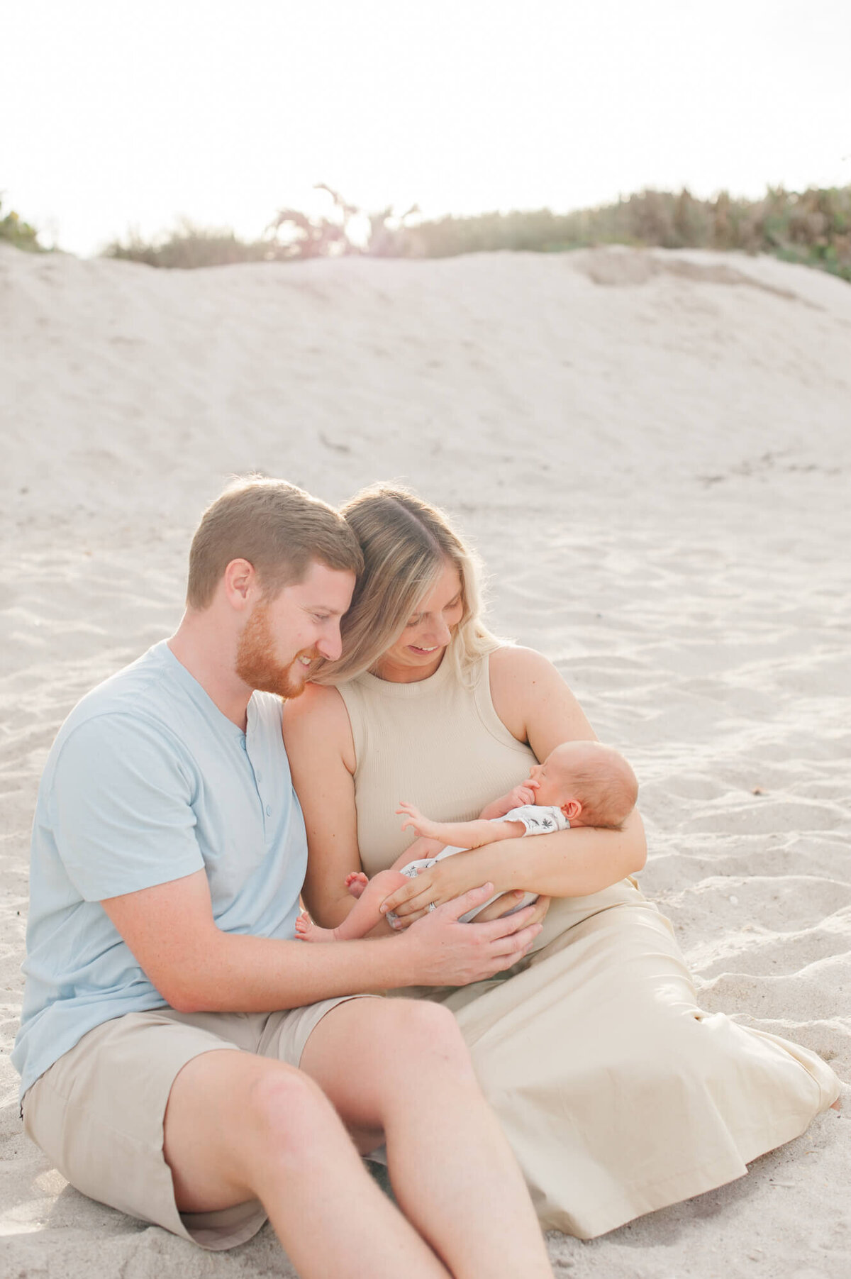 New parents sit in the sand holding their newborn son at golden hour