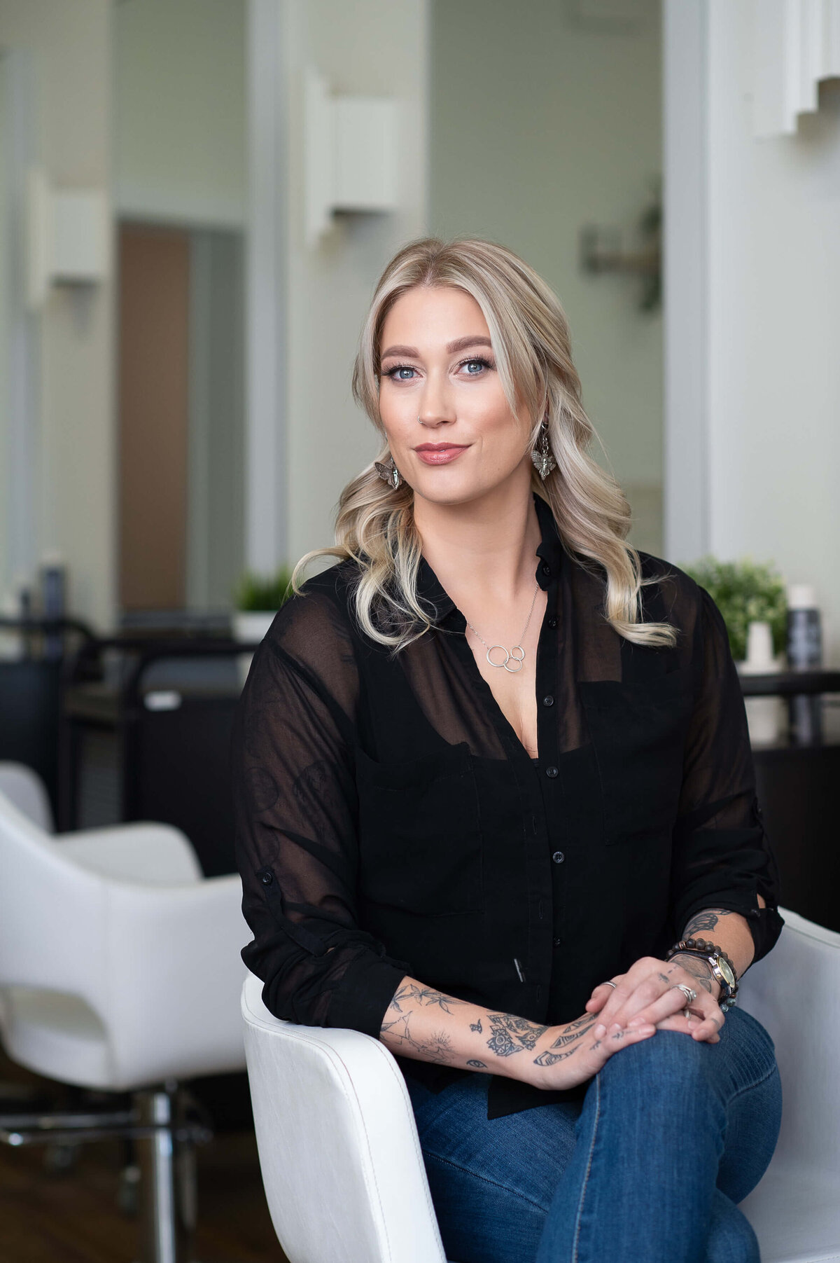 a profile photo of a blonde hair stylist in a black top and jeans sitting in a white chair.  Captured by Ottawa branding photographer JEMMAN Photography