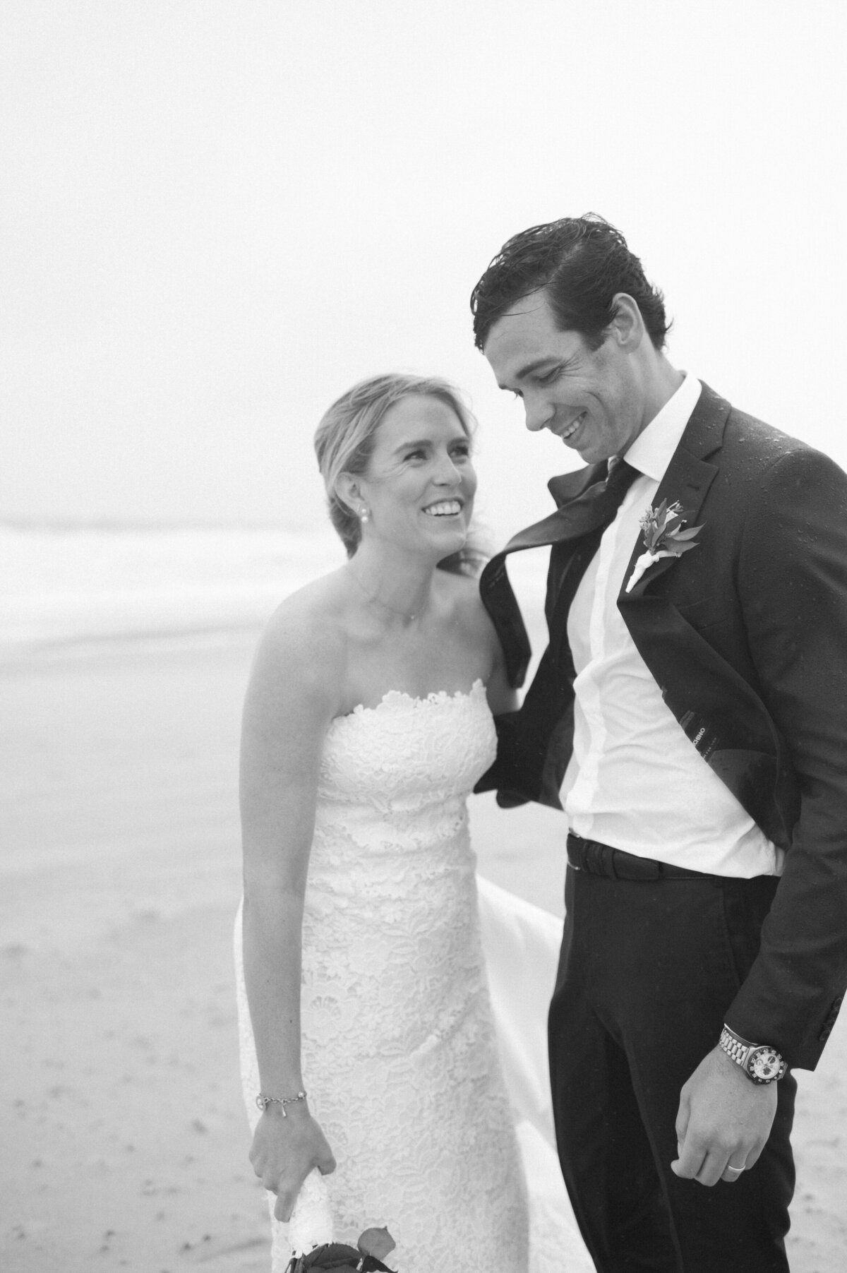 The couple stands close, smiling at each other after braving the rain on the beach