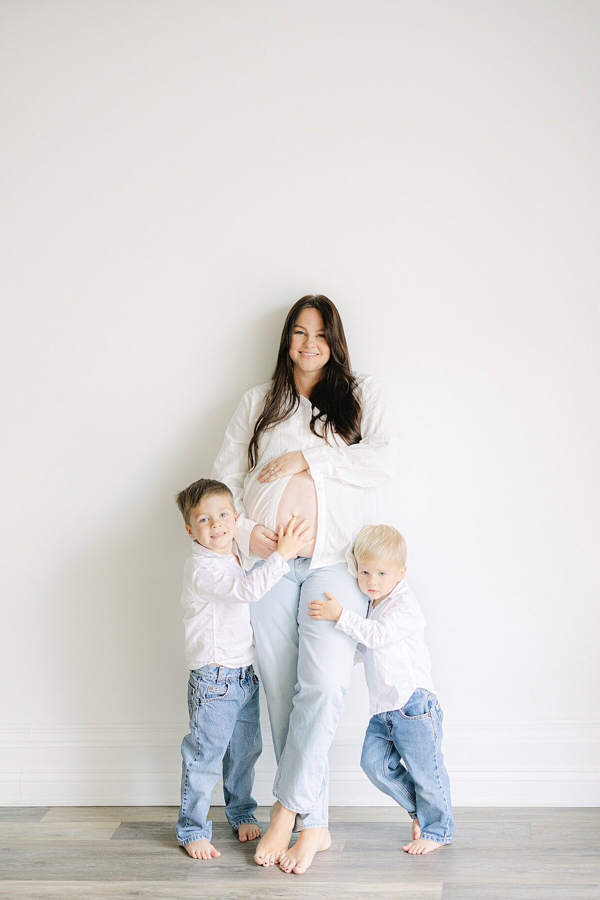Pregnant mother and her two young boys dressed in jeans and white button down tops smile at Indianapolis family photographer, Brittney Lear
