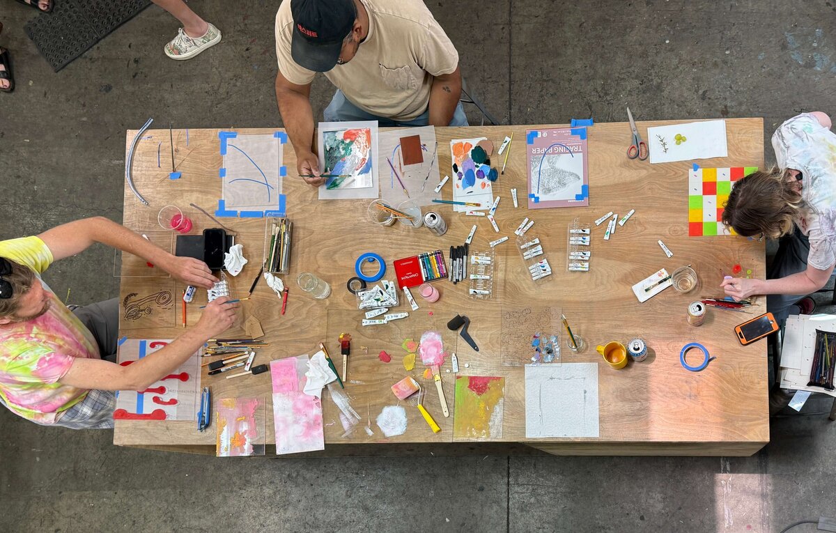 Overhead view of multiple artists at a large table working on their monoprints using vibrant pigments, tools, and layered plexiglass plates.